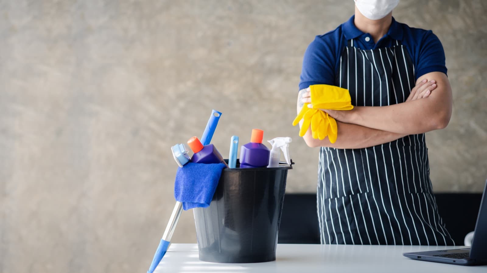 Cleaning professional in a striped apron standing with arms crossed next to a bucket of supplies
