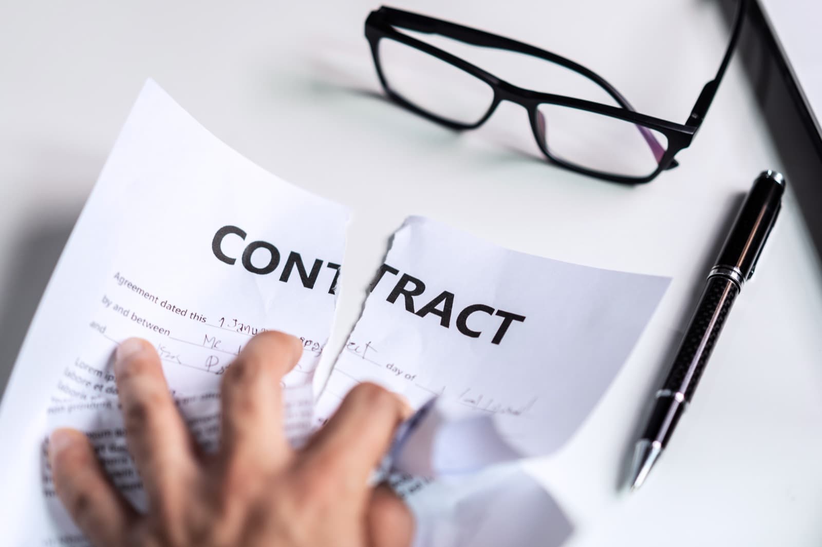 Hand tearing a contract document in half on a desk with glasses and a pen